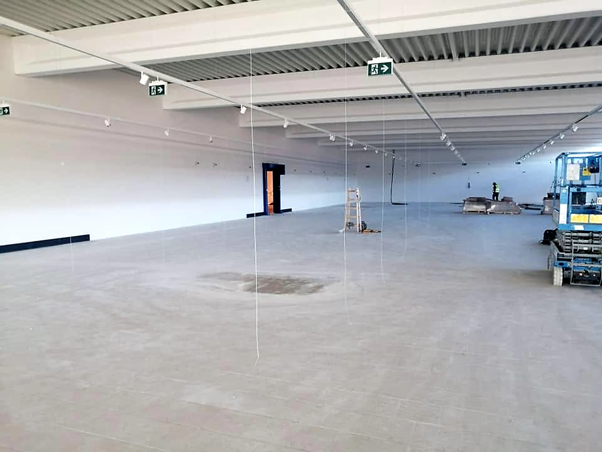 Duża sala sprzedaży z pomalowanym stropem z blachy trapezowej, szynami oświetleniowymi i znakami wyjścia ewakuacyjnego; z prawej podnośnik nożycowy