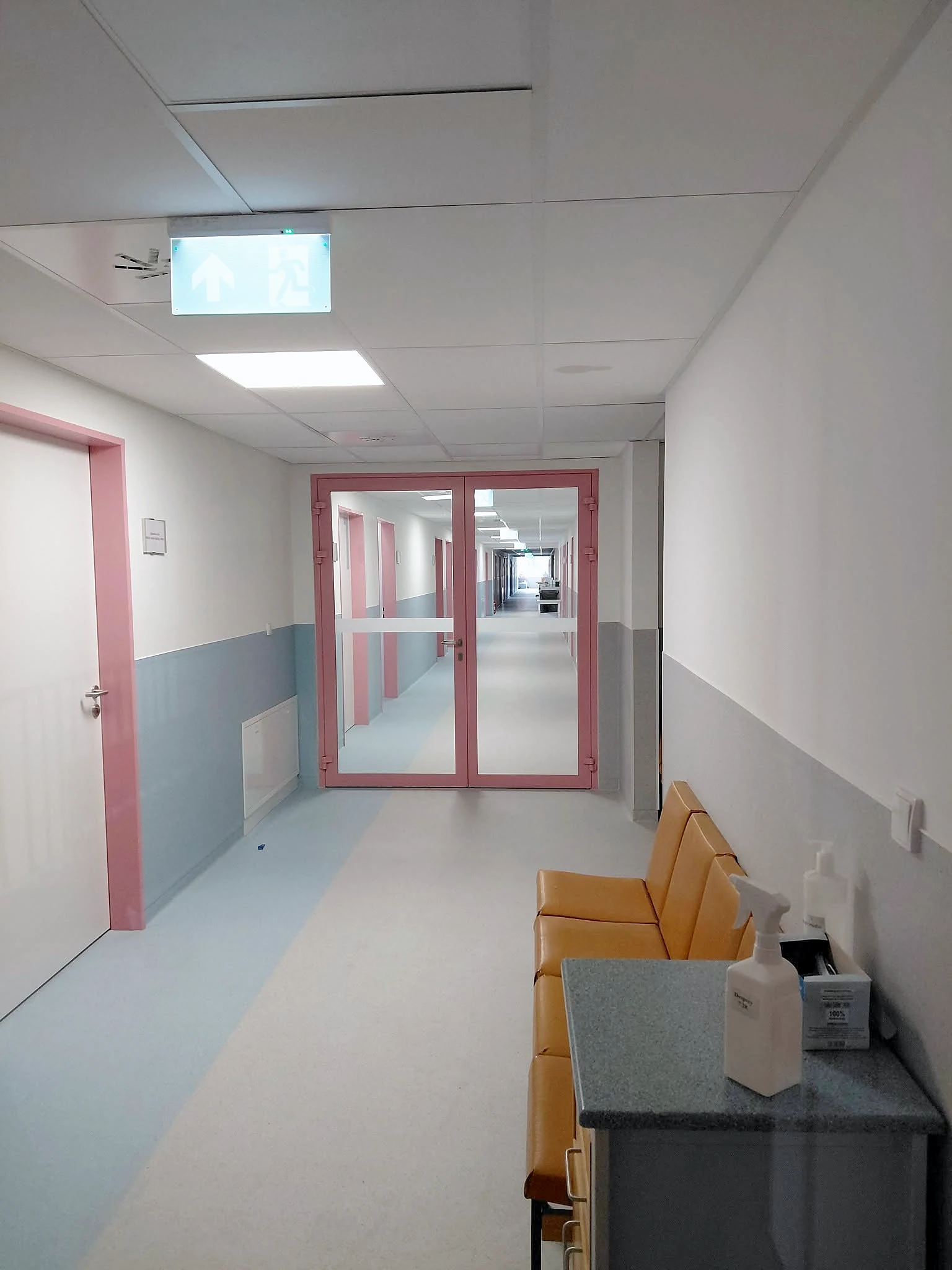 Wykończony korytarz przychodni: różowe drzwi przeciwpożarowe, sufit kasetonowy, krzesła w poczekalni